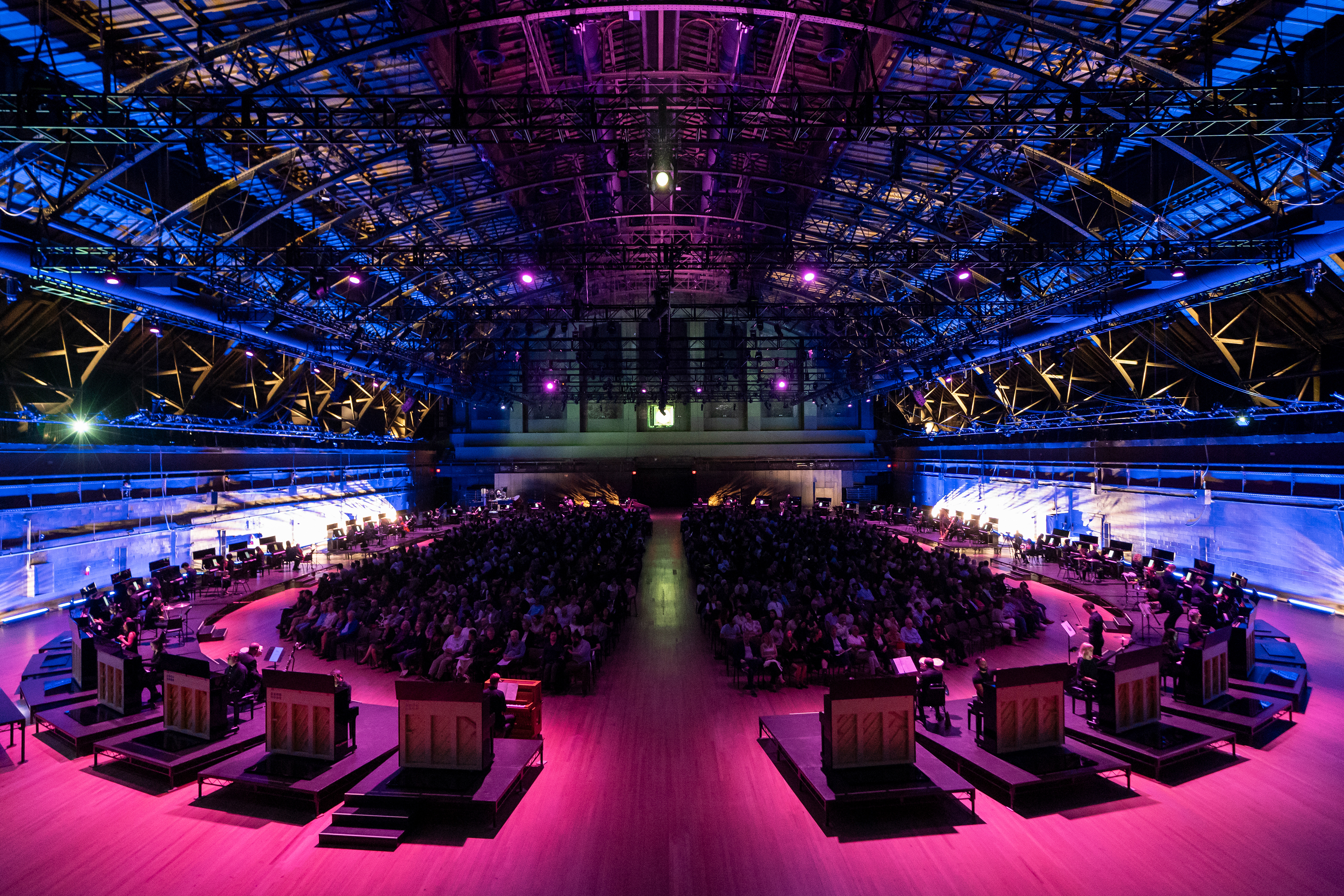 50 pianos encircle an audience of hundreds of concert-goers, bathed in fuchsia and blue lighting.