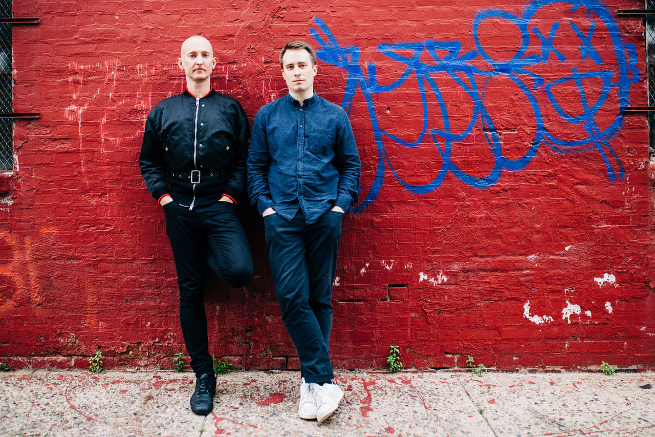 Philip Venables and Ted Huffman stand together in front of a building with a bold red brick wall with blue graffiti marks.