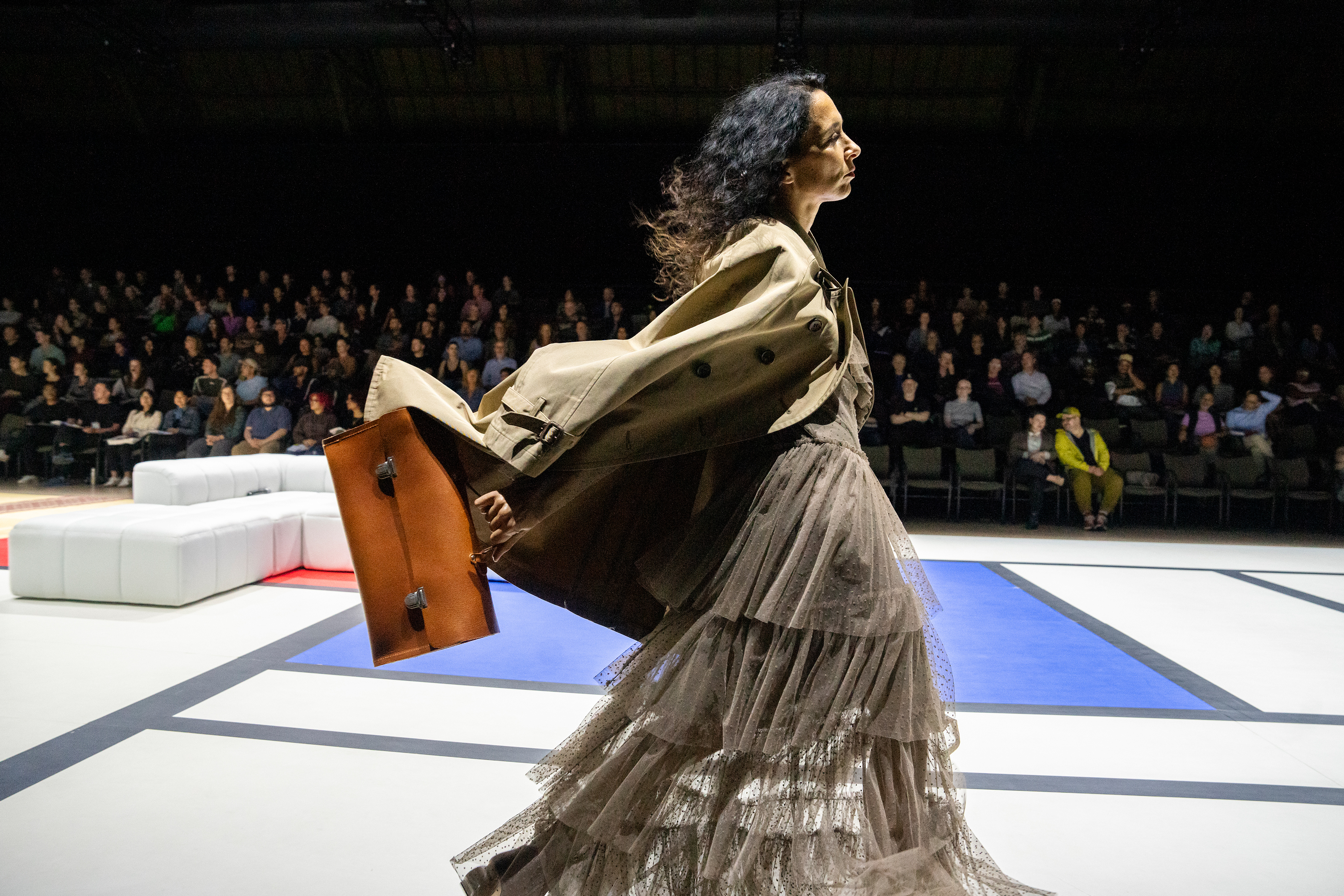 A fashionable dancer carrying a briefcase walks along a red, white, and blue dance floor in front of a crowd of spectators.
