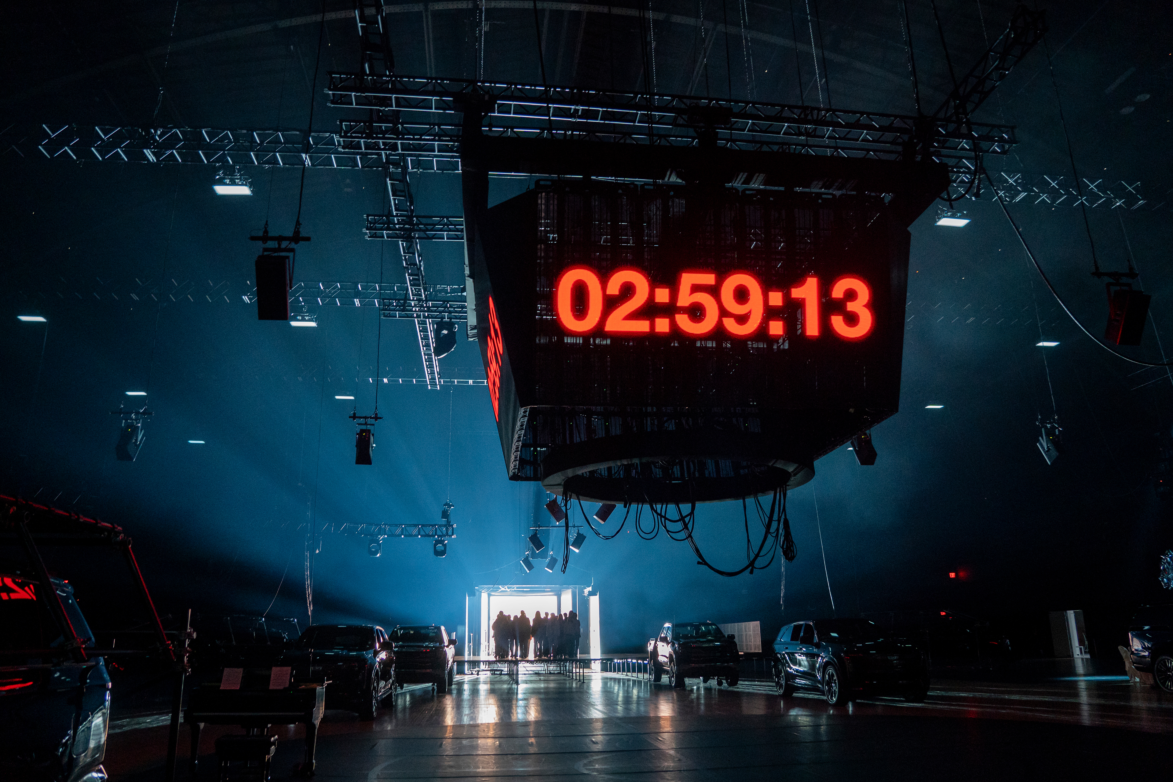 A group of performers enters the performance space, which includes a countdown clock on a Jumbo-Tron and Cadillac Escalades.